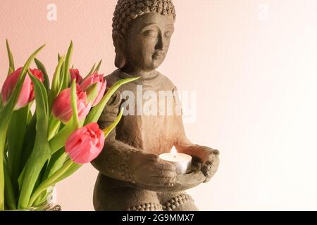 Kleine buddha-Skulptur aus Keramik mit brennender Kerze und rosa Tulpen vor hellem Hintergrund mit Kopierraum, ausgesuchter Weichfokus, schmal Stockfoto