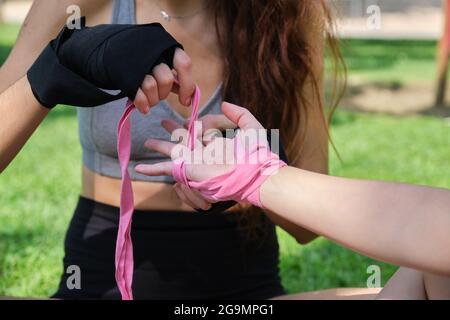Eine nicht erkennbare Person, die anderen hilft, muay thai-Handpackungen anzuziehen, die in einem Park auf Gras sitzen. Sport im Freien. Stockfoto