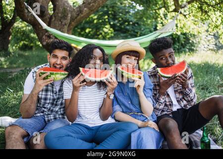 Eine Gruppe von vier multikulturellen Menschen, die süße reife Wassermelone essen, während sie im grünen Garten picknicken. Glückliche Freunde, die gemeinsam Freizeit verbringen. Entspannung in der Natur. Stockfoto