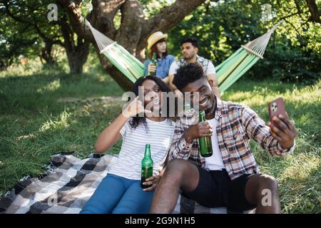 Lustiger schwarzer Mann und Frau, die sich im Sommerpark entspannen und Selfie auf dem Handy machen. Verwischen Sie den Hintergrund zweier verschiedener Freunde, die in der Hängematte mit Bier in den Händen sitzen. Stockfoto