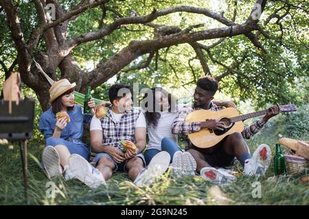 Sorglose, vielfältige Menschen sitzen im Sommergarten mit Fast Food und Trinken und spielen Gitarre. Vier multiethnische Freunde verbringen ihre Wochenenden mit Vergnügen in der Natur. Stockfoto