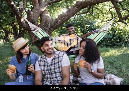 afroamerikanischer Kerl, der in der Hängematte sitzt und Gitarre spielt, während seine drei multikulturellen Freunde sich auf dem Gras mit Getränken und Snacks entspannen. Konzept von Mensch, Freizeit und Natur. Stockfoto