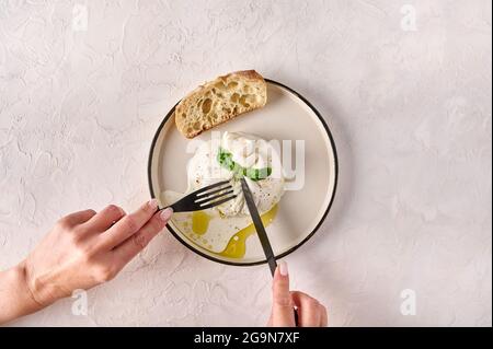 Damenhänden mit Messer und Gabel Burrata Käse mit Basilikum, Öl und Brot auf weißem Teller geschnitten. Draufsicht. Speicherplatz kopieren Stockfoto