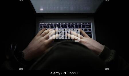 Ein einfaches, sehr dunkles Nachtbild mit Händen auf einer beleuchteten Tastatur, die tippt. Schattige Person, die im Dunkeln an einem Computer oder Laptop eine Kapuze trägt. Stockfoto