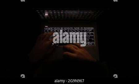 Ein einfaches, sehr dunkles Nachtbild mit Händen auf einer beleuchteten Tastatur, die tippt. Computer oder Laptop im Dunkeln verwenden. Stockfoto