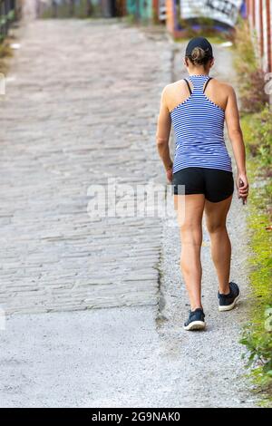 Junge Frau, die in der Sommersonne Shorts und Weste mit Baseballmütze trägt, geht auf einer gepflasterten Straße entlang, um sich fit zu halten. Stockfoto