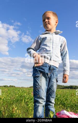 Ein zufriedenes Vorschulkind in Jeans und einem Pullover hält Papiergeld in der Natur auf einem Feld vor dem Hintergrund eines blauen Himmels mit Wolken. Hochformat Stockfoto