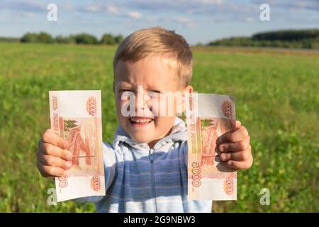 Ein zufriedenes Kind, ein Vorschulkind, hält Papierrubel in seinen Händen draußen auf dem Hintergrund eines Feldes mit grünem Gras an einem hellen sonnigen Sommer Stockfoto