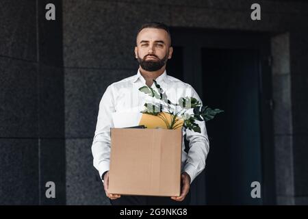 Arbeitsloser Mann, der mit einer Schachtel voller persönlicher Sachen aus dem Bürozentrum auszieht und traurig vor die Kamera schaut Stockfoto