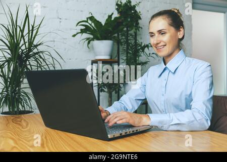 Lächelnde Frau, die in einem hellen, modernen Büro mit grünen Pflanzen am Laptop arbeitet Stockfoto