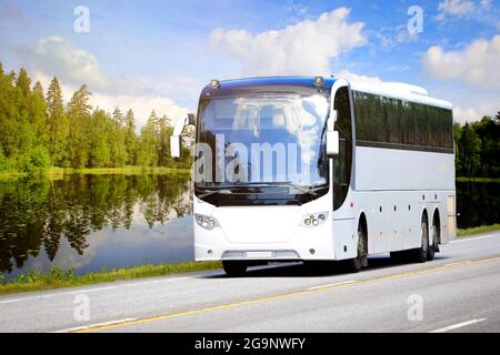 Weißer Reisebus fährt auf der Autobahn durch die ländliche Landschaft eines ruhigen Sees, grünen Waldes und blauen Himmels mit fairweather-Wolken an einem Sommertag. Stockfoto