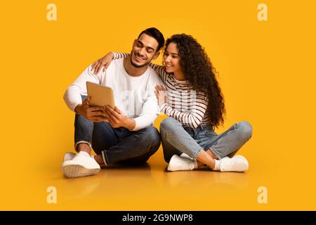 Happy Young Middle-Eastern Paar Mit Digital Tablet, Während Sie Über Gelben Hintergrund Sitzen Stockfoto
