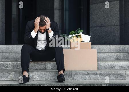 Verärgert reifer Geschäftsmann sitzt mit leerem Plakat Pappschild und Box mit persönlichen Sachen auf der Treppe des Bürozentrums Stockfoto