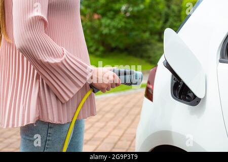 Frau, die einen Ladestecker oder ein Ladegerät in ein weißes Elektroauto steckt Stockfoto