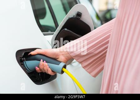 Weibliche Hand, öffnen Sie eine Steckdosenabdeckung und stecken Sie das Ladegerät in ein Elektroauto an einer Ladestation Stockfoto