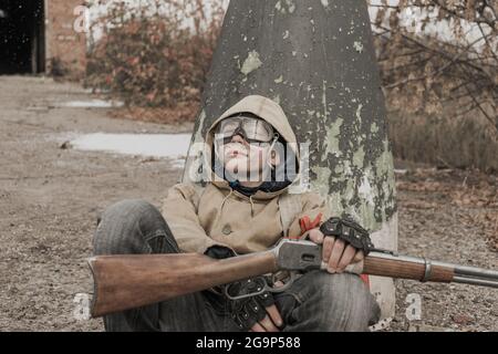 Wandernder Junge. Junge mit einer Waffe. Junge geht zu einem verlassenen Gebäude. Junge steht vor einem Gebäude. Post-Apokalypse. Mann, der zu Fuß in einem Stockfoto