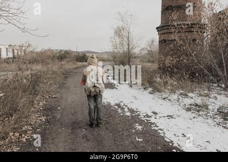 Wandernder Junge. Junge mit einer Waffe. Junge geht zu einem verlassenen Gebäude. Junge steht vor einem Gebäude. Post-Apokalypse. Mann, der zu Fuß in einem Stockfoto