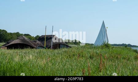 Strohgedeckte Bootshäuser schmiegen sich an die hohen Grashalme auf Hickling Broad, östlich von Norwich, in Norfolk UK. Segelboot in der Ferne. Stockfoto