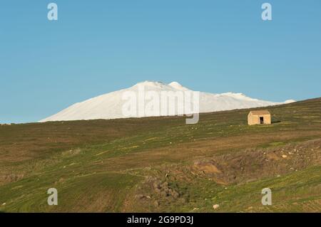 Grüner Hang mit einer alten Hütte und weißem Ätna, der gegen den klaren Himmel des Abends schneebedeckt ist Stockfoto