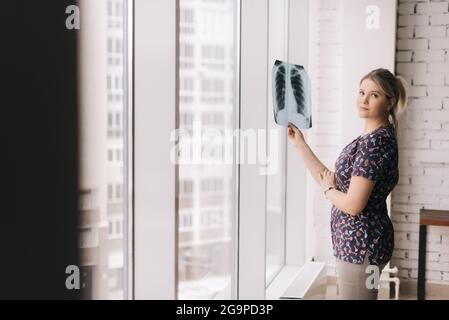 Selbstbewusste junge Frau, die an sonnigen Tagen ein Röntgenbild der Lunge am Fenster hält und die Kamera anschaut. Stockfoto