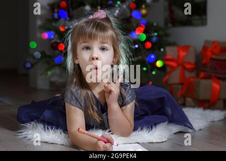 Kindermädchen in einem blauen Kleid begeistert schreibt einen Brief an Santa durch den Weihnachtsbaum. Baby hob ihren Daumen nach oben, Konzept der Idee Stockfoto