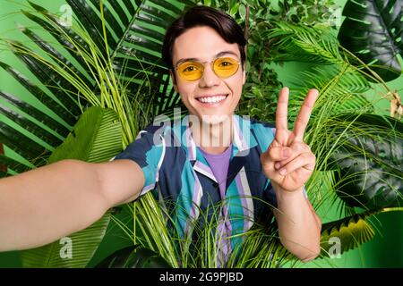 Foto von lustigen Brunet Millennial Kerl im Dschungel tun Selfie V-Zeichen tragen Brille Hemd isoliert auf grünem Hintergrund Stockfoto