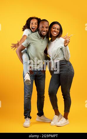 Fröhliche afroamerikanische Familie mit niedlichen Tochter posiert über gelben Hintergrund, Spaß, Mädchen Huckepack Papa Stockfoto