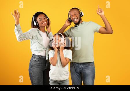 Genießen Sie Musik zusammen. Glückliche afroamerikanische Dreierfamilie, die in Kopfhörern tanzt, isoliert auf gelbem Hintergrund Stockfoto