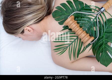 Madero-Therapie, Entspannungsmassage - Frau, die eine Spa-Rückenmassage mit einem Holzrollermassager macht, das auf grünen Blättern auf ihrem Rücken liegt, Nahaufnahme, Draufsicht. Stockfoto