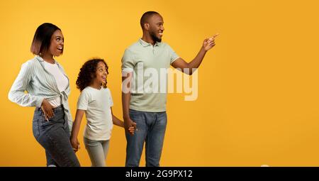 Positive afroamerikanische Dreierfamilie schaut beiseite, junger Mann zeigt mit dem Finger auf den Kopierraum auf gelbem Hintergrund Stockfoto