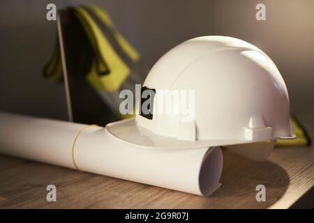 Weißer Schutzhelm, gerollte Blaupläne, Sicherheitsweste und Laptop-Computer auf dem Tisch des Technikers. Bau-, Design-, Architektur- oder Bauindustriekonzept. Hochwertige Fotos Stockfoto