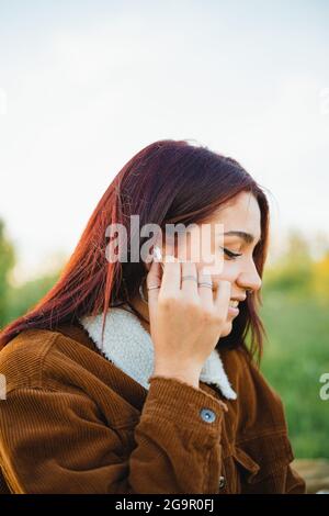Ein Teenager, der ein weißes schnurloses Ohrtelefon in ihr Ohr setzt, während er während des Sonnenuntergangs auf dem Gras sitzt Stockfoto
