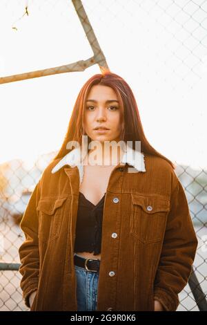 Porträt eines hübschen Teenagers, das gegen einen Kettenzaun steht und während des Sonnenuntergangs die Kamera anschaut. Sie trägt eine braune Jacke und blaue Jeans. Stockfoto