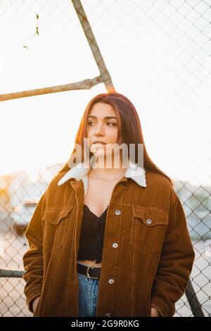 Porträt eines hübschen Teenagers, das während des Sonnenuntergangs gegen einen Kettenzaun steht. Sie trägt eine braune Jacke und blaue Jeans. Stockfoto