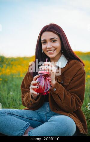 Lächelndes Teenager-Mädchen, das Wasser in einem roten Glas trinkt, das auf dem Gras eines Hügels sitzt. Sie schaut in die Kamera. Stockfoto