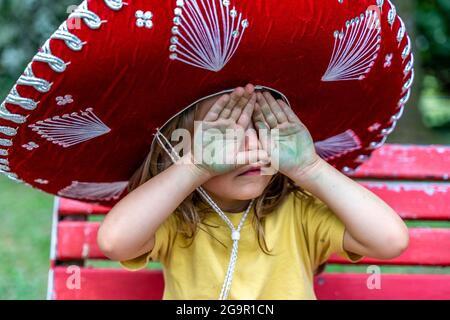 Das kleine süße Mädchen, das einen Sombrero trägt, bedeckt ihre Augen mit grünen schmutzigen Händen Stockfoto