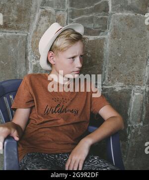 Teenager 10 Jahre alt, kaukasische Nationalität in einem Strohhut, blond, sitzt auf einem Stuhl vor dem Hintergrund einer Steinmauer.moderner Junge. Stockfoto