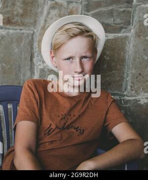 Teenager 10 Jahre alt, kaukasische Nationalität in einem Strohhut, blond, sitzt auf einem Stuhl vor dem Hintergrund einer Steinmauer.moderner Junge. Stockfoto