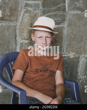 Teenager 10 Jahre alt, kaukasische Nationalität in einem Strohhut, blond, sitzt auf einem Stuhl vor dem Hintergrund einer Steinmauer.moderner Junge. Stockfoto