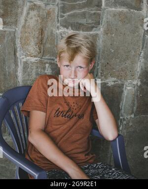 Teenager 10 Jahre alt, kaukasische Nationalität in einem Strohhut, blond, sitzt auf einem Stuhl vor dem Hintergrund einer Steinmauer.moderner Junge. Stockfoto