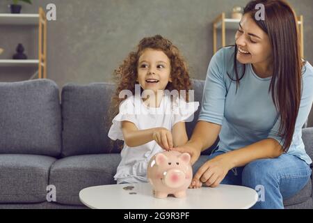 Glückliche junge Mutter und kleine Tochter, die zusammen Münzen in ihre Sparschwein legen Stockfoto