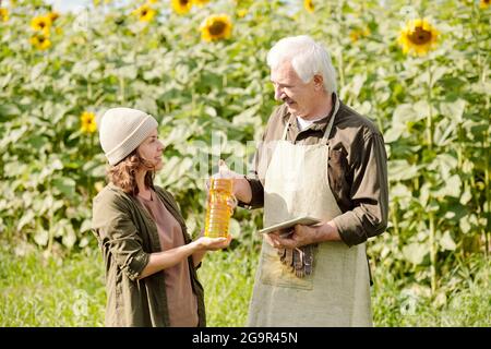 Happy Senior männlichen Landwirt in Arbeitskleidung vorbei Flasche Sonnenblume Öl an seine reife Kollegin gegen grünes Feld an Sonniger Sommertag Stockfoto