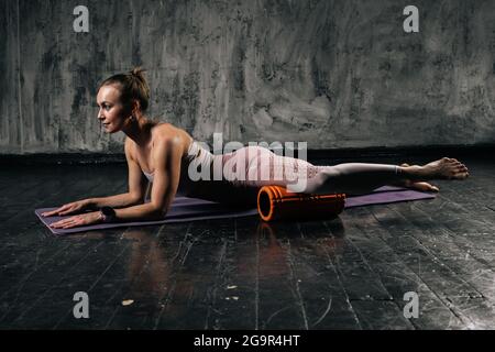 Muskulöse junge athletische Frau mit perfektem, schönem Körper im aktiven Ohr massiert die Fußmuskeln mit dem Schaumstoffroller-Massagegerät, das auf einer Yogamatte liegt. Stockfoto