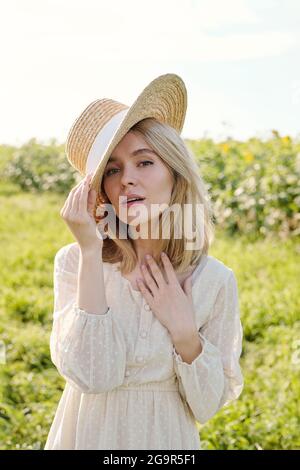 Wunderschöne junge blonde Frau in Strohhut und weißen Land Style-Kleid vor der Kamera gegen grünes Gras stehen Und Sonnenblumenfeld Stockfoto