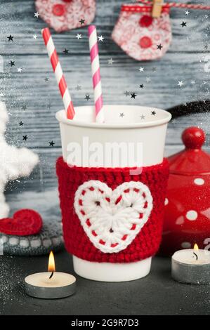 Becher mit Trinkhalmen in rot gestricktem Wärmer auf blauem Holzhintergrund.Weihnachtsset mit Teekannen und brennenden Kerzen Stockfoto