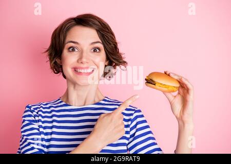 Porträt von jungen ekstatischen attraktiven fröhlich lächelnd Mädchen zeigt Finger auf Cheeseburger isoliert auf rosa Hintergrund Stockfoto