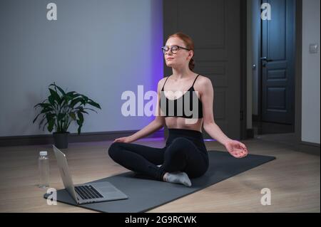 Während der Quarantäne führt das süße Mädchen einen gesunden Lebensstil, sie macht online Yoga, sitzt in der Lotusposition vor dem Laptop. Stockfoto