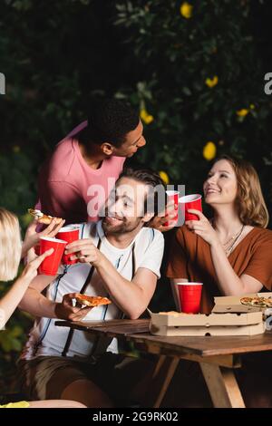 Multiethnische Freunde mit Pizza und Plastikbechern reden während der Party Stockfoto