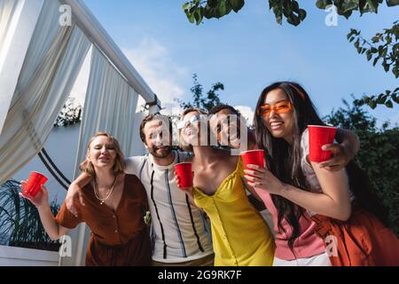 Fröhliche interracial junge Menschen mit Plastikbechern umarmen im Freien Stockfoto