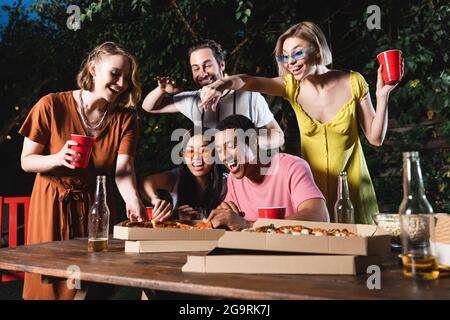 Glückliche interracial Freunde mit Plastikbechern sitzen in der Nähe von Pizza im Freien Stockfoto